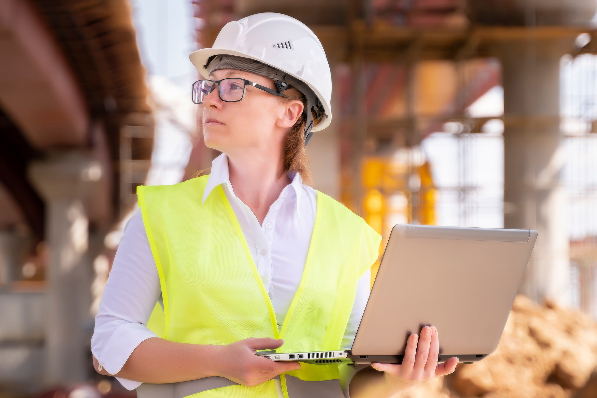 Eine Vorarbeiterin mit weißem Helm und gelber Weste steht mit einem Laptop in der Hand auf einer Baustelle.