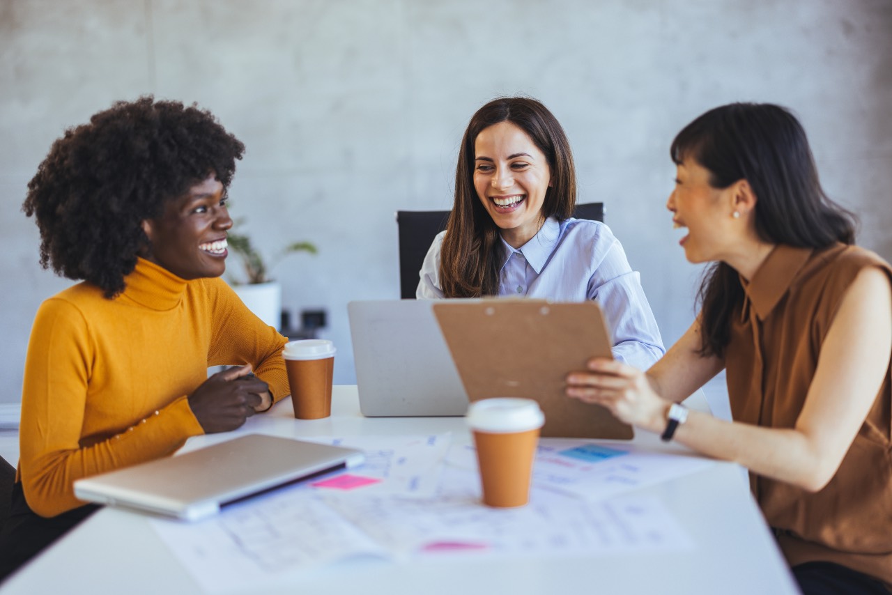Drei_Frauen_unterschiedlicher_Herkunft Drei Frauen unterschiedlicher Herkunft sitzen an einem Tisch mit einem Laptop und lachen.