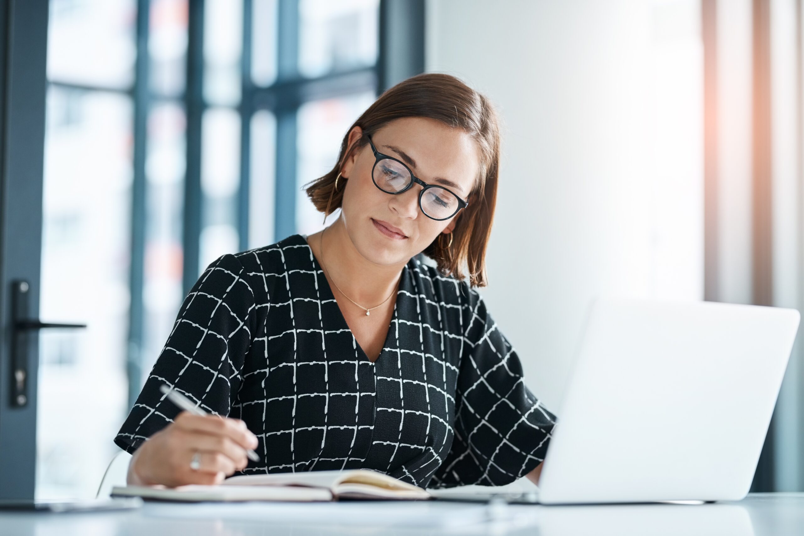 Office,,Woman,And,Laptop,Or,Writing,In,Journal,For,Proposal, Frau mit Brille am Laptop. Sie schreibt gerade etwas mit einem Stift in der Hand.