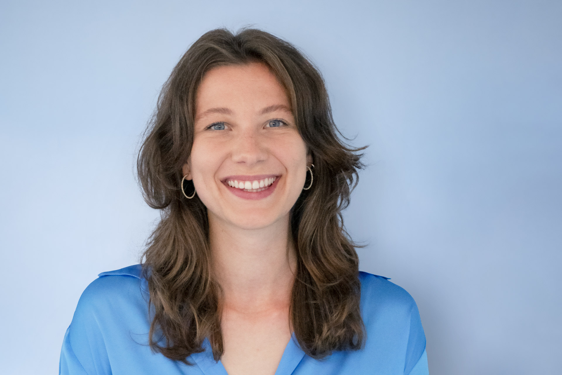 Nina_Voessing_Artikel Lächelnde Frau mit blauer Bluse und braunen Haaren. Hellblauer Hintergrund