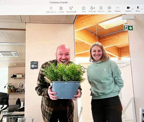 Julien bekommt vom Team ein Geschenk überreicht :) Ein Mann und eine Frau stehen lächelnd vor eine Webcam. Der Mann hält begeistert einen Topf mit verschiedenen Kräutern hoch.